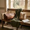 Bolsa de viaje de cuero marrón y abrigo ligero preparados sobre un banco de madera junto a una ventana luminosa