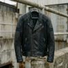 Cazadora moto de cuero negro con paneles técnicos colgada sobre un fondo urbano de hormigón con luz natural.