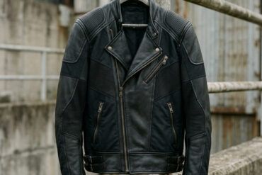 Cazadora moto de cuero negro con paneles técnicos colgada sobre un fondo urbano de hormigón con luz natural.