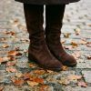 Detalle de botas altas de serraje marrón chocolate sobre hojas secas de otoño y pavimento de piedra