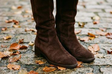 Detalle de botas altas de serraje marrón chocolate sobre hojas secas de otoño y pavimento de piedra