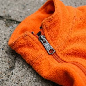 Detalle de tejido técnico naranja de alta visibilidad con cremallera sobre fondo de asfalto urbano.