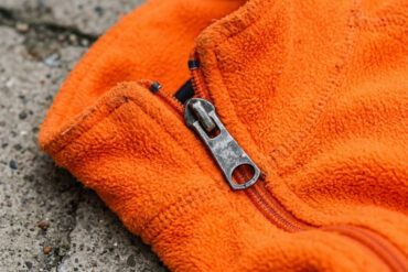 Detalle de tejido técnico naranja de alta visibilidad con cremallera sobre fondo de asfalto urbano.