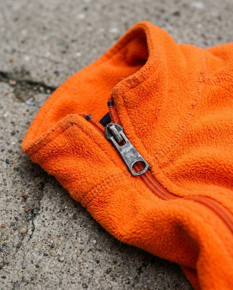 Detalle de tejido técnico naranja de alta visibilidad con cremallera sobre fondo de asfalto urbano.