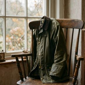 Chaqueta encerada verde oliva con cuello de pana apoyada en una silla de madera junto a una ventana con gotas de lluvia.