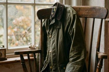 Chaqueta encerada verde oliva con cuello de pana apoyada en una silla de madera junto a una ventana con gotas de lluvia.