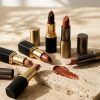 Tres barras de labiales marrones en tonos tostados y chocolate colocadas sobre una bandeja minimalista con luz natural.