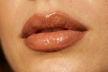 Primer plano de labios carnosos con perfilado cálido y acabado brillante de aceite labial con luz natural.