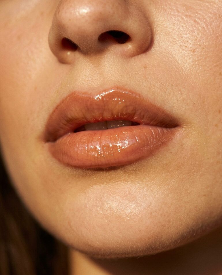 Primer plano de labios carnosos con perfilado cálido y acabado brillante de aceite labial con luz natural.