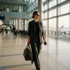 Mujer con traje sastre sofisticado y bolso de viaje caminando por un espacio arquitectónico luminoso.