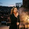 Cantante sobre el escenario con micrófono bajo iluminación cálida en un anfiteatro nocturno con público de fondo
