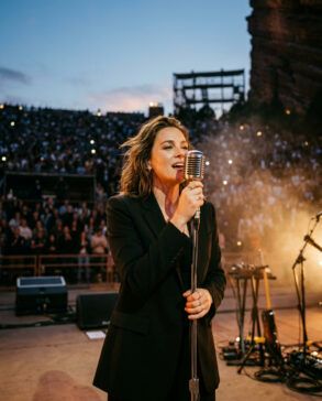 Cantante sobre el escenario con micrófono bajo iluminación cálida en un anfiteatro nocturno con público de fondo