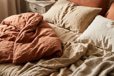 Primer plano de sábanas de lino arrugadas en tono terracota y arena sobre una cama iluminada con luz natural matutina