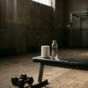 Bote de suplemento deportivo junto a una botella de agua y mancuernas en un gimnasio iluminado por luz matinal.