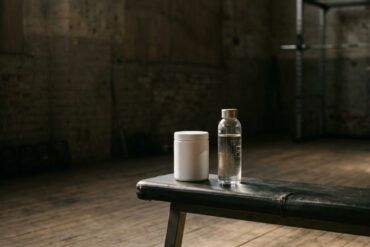 Bote de suplemento deportivo junto a una botella de agua y mancuernas en un gimnasio iluminado por luz matinal.