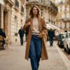Mujer vestida con vaqueros de tiro bajo de corte recto, jersey de punto fino y abrigo largo en una calle otoñal.